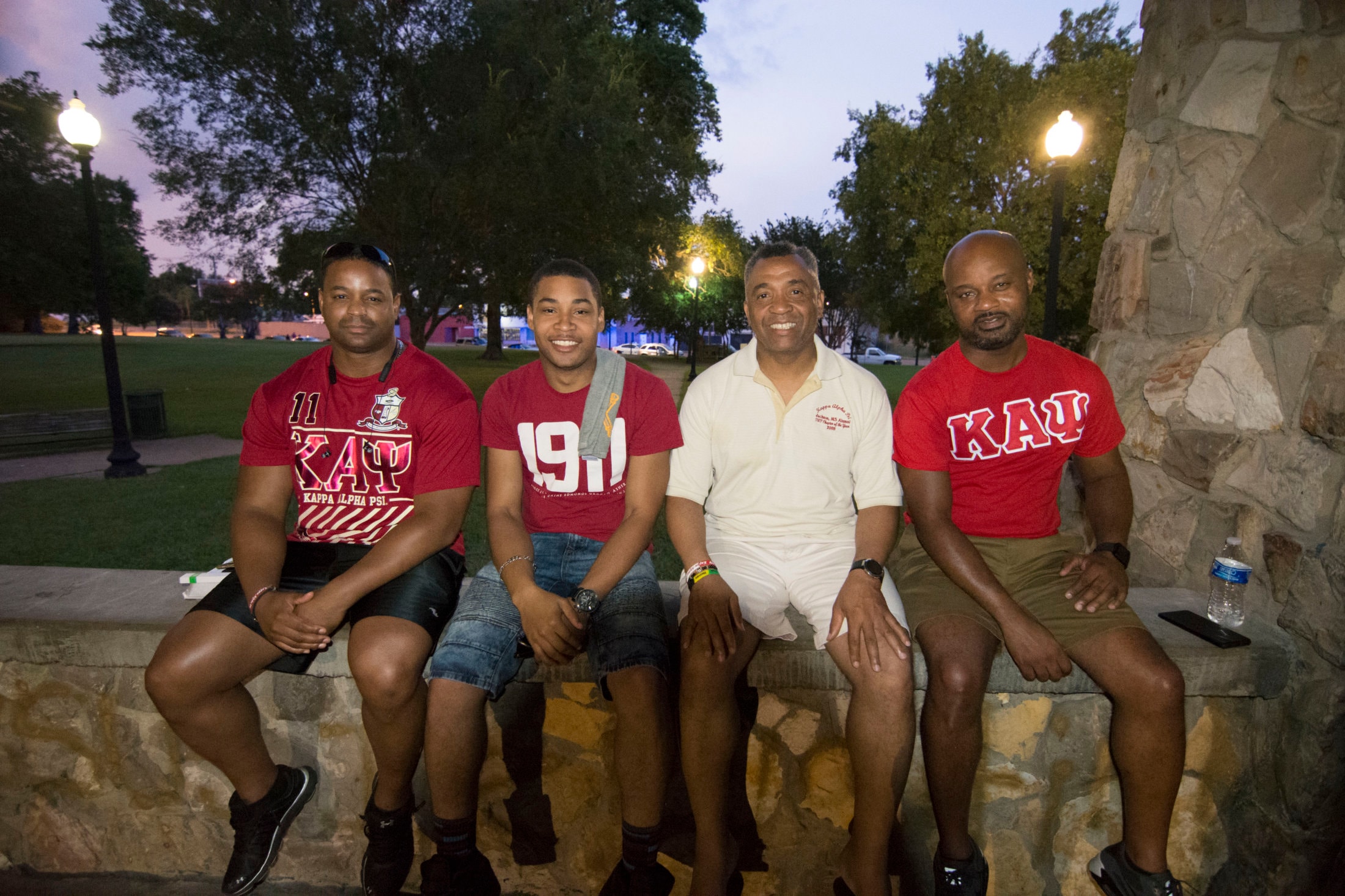 Brothers in Kappa Alpha Psi shirts seated on stone wall at dusk