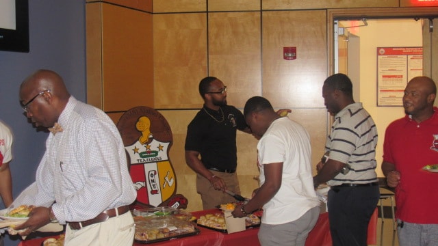 Brothers at chapter gathering with Kappa Alpha Psi shield on wall